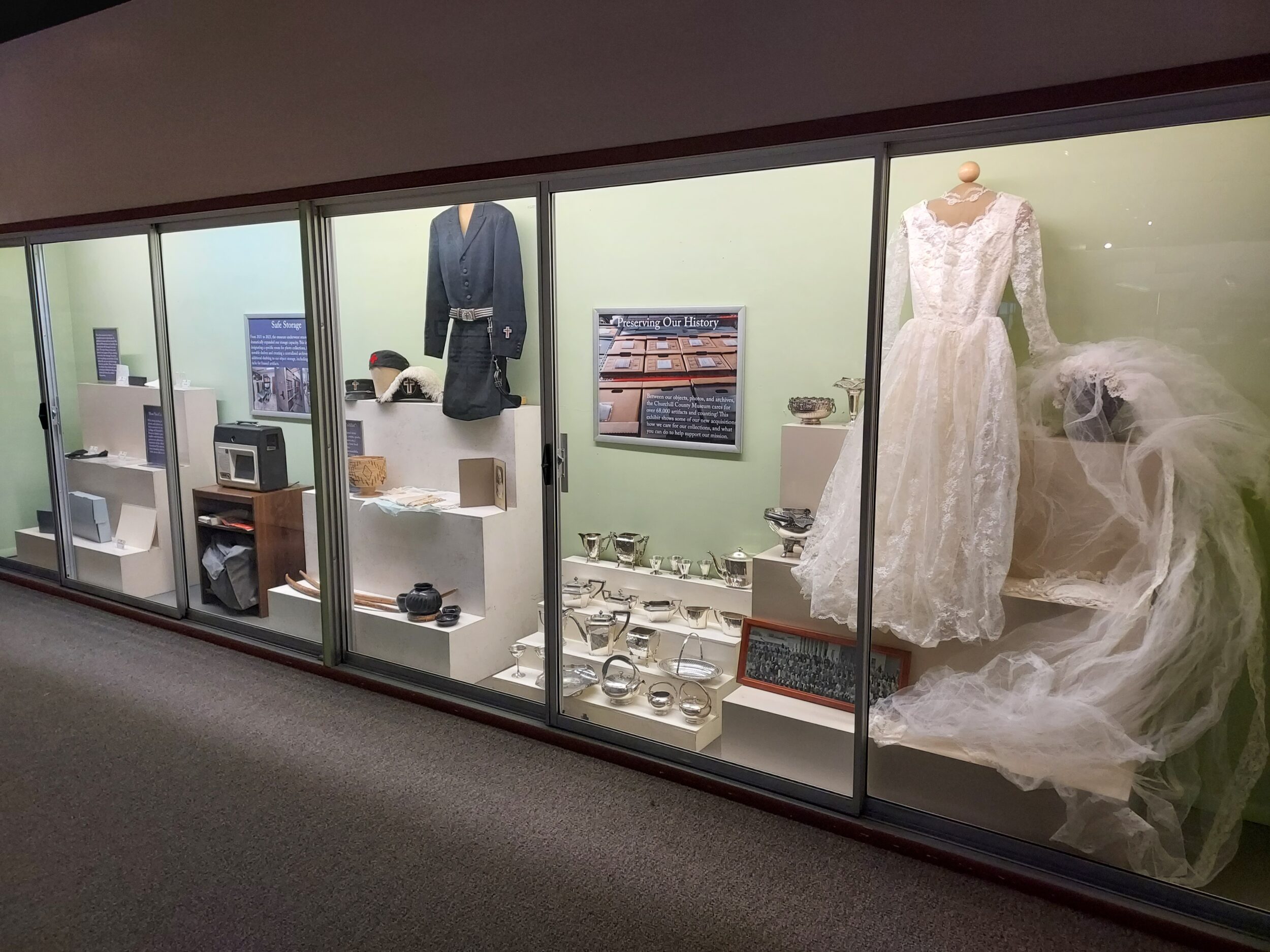 [A photo of a long green museum case with several white tiered display stands holding various artifacts, along with storage supplies such as boxes, folders, and photo sleeves]