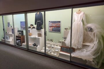[A photo of a long green museum case with several white tiered display stands holding various artifacts, along with storage supplies such as boxes, folders, and photo sleeves]